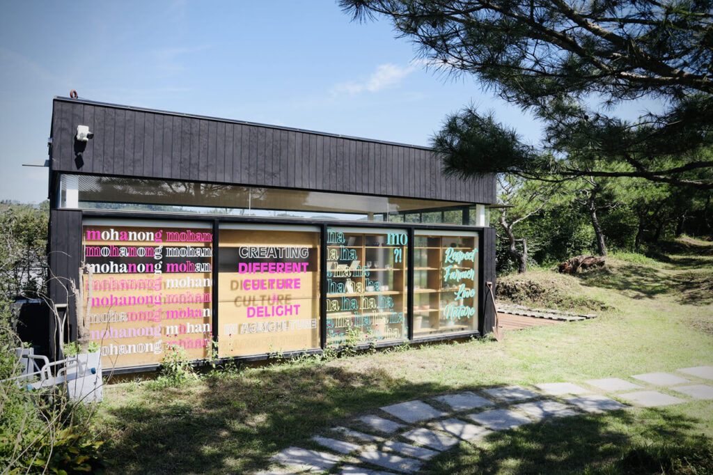 녹색 잔디밭 위에 있는 검은색 세로 결 목재로 이루어진 벽체와 지붕, 그리고 통유리 벽으로 구성된 뭐하농 하우스 (Mohanong house) 의 물품 매장 건물