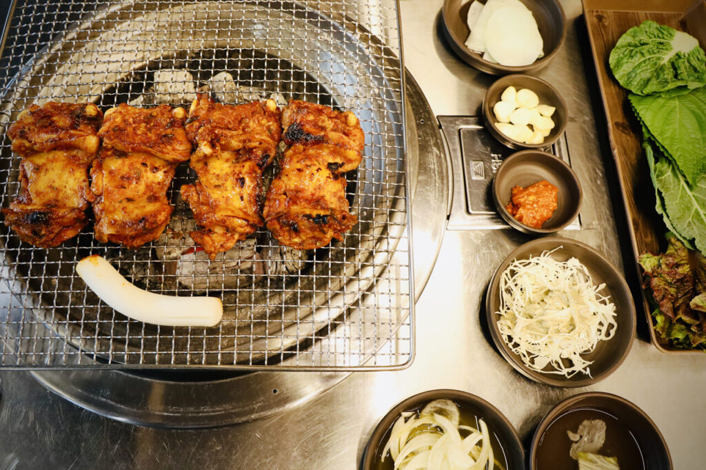 숯불 위 은색 철망에 올려진 샘토 명물 닭갈비의 붉은빛 양념 닭갈비 네 덩어리와 그 옆에 삼무, 마늘, 쌈장, 샐러드 녹색 쌈 채소가 놓인 모습