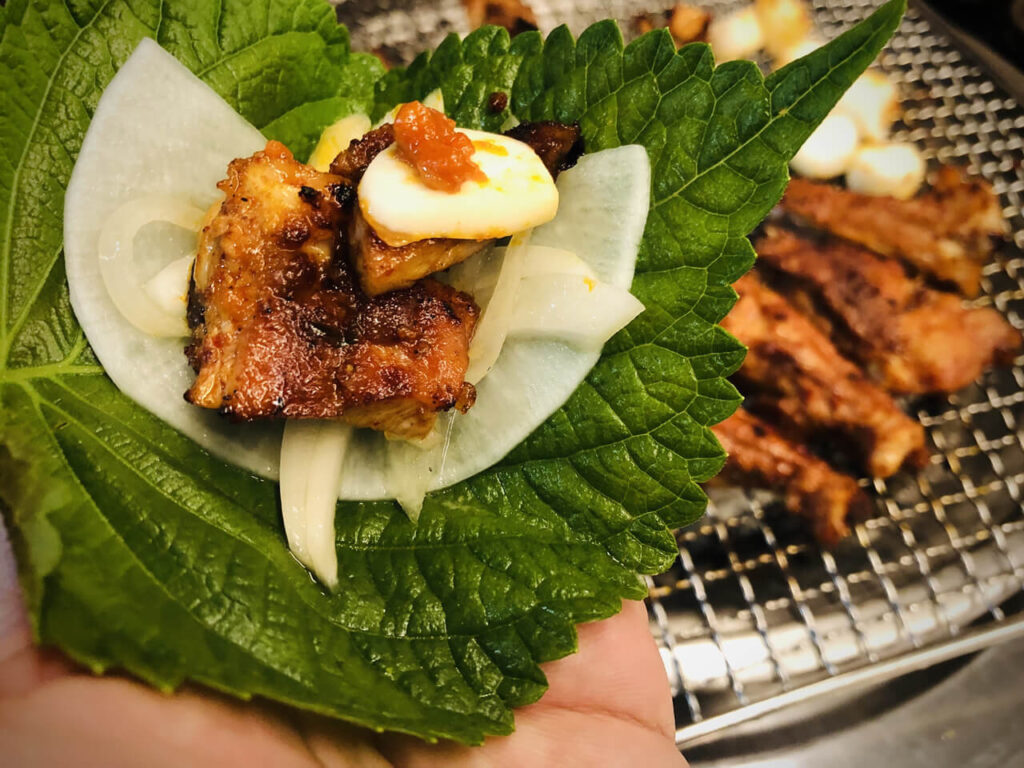 녹색 깻잎 위에 흰색 쌈무와 그 위에 올려진 샘토 명물 닭갈비의 양념 닭갈비 한 조각, 그리고 쌈장 묻힌 생마늘